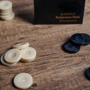 Puede incluir: Un juego de backgammon con fichas blancas y negras. Las fichas blancas est&aacute;n apiladas, mientras que las fichas negras est&aacute;n esparcidas sobre una superficie de madera. La caja del juego est&aacute; en el fondo, con el texto "Galaith Backgammon Dice Exclusive" impreso en la parte superior.