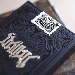 Op de afbeelding: Een zwart deck speelkaarten met een zilveren folieontwerp en de tekst "The Stranger" en een kleine zwart-witte stempel met een ontwerp.