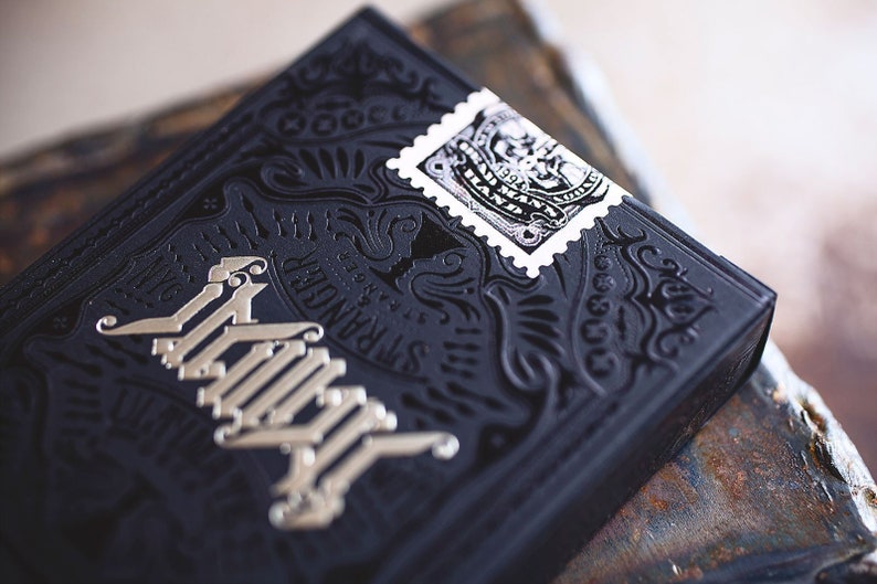 Op de afbeelding: Een zwart deck speelkaarten met een zilveren folieontwerp en de tekst "The Stranger" en een kleine zwart-witte stempel met een ontwerp.