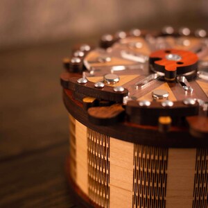 Puede incluir: Una caja de rompecabezas de madera con una tapa circular y un dise&ntilde;o geom&eacute;trico. La caja est&aacute; hecha de madera clara y oscura y tiene un s&iacute;mbolo yin yang rojo y blanco en la parte superior.