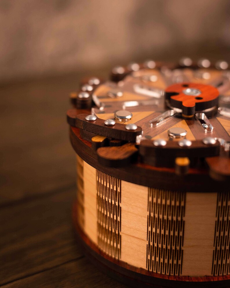 Puede incluir: Una caja de rompecabezas de madera con una tapa circular y un dise&ntilde;o geom&eacute;trico. La caja est&aacute; hecha de madera clara y oscura y tiene un s&iacute;mbolo yin yang rojo y blanco en la parte superior.