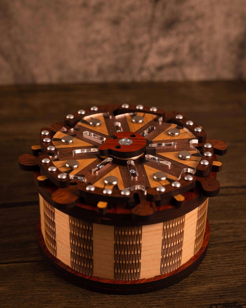 Puede incluir: Una caja de rompecabezas de madera con una tapa circular y un dise&ntilde;o intrincado. La tapa est&aacute; hecha de m&uacute;ltiples piezas de madera, cada una con un patr&oacute;n de veta diferente. La caja est&aacute; cerrada y la tapa est&aacute; ligeramente levantada.