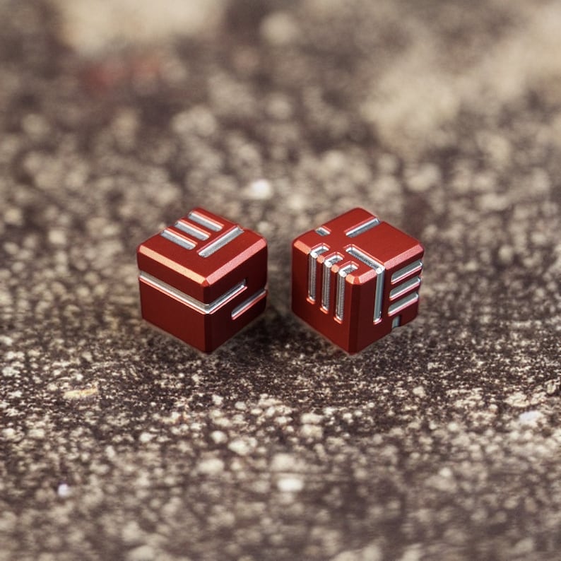 Op de afbeelding: Twee kleine, rode, kubusvormige objecten met zilveren accenten. Elke kubus heeft een uniek geometrisch ontwerp op het oppervlak. De objecten liggen op een gestructureerd, grijs oppervlak.