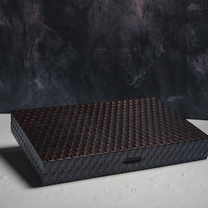 以下が含まれることがあります： 暗い茶色と赤色の織り目のような模様が施された、長方形の黒い木製の箱。 箱には小さな長方形の留め金があり、暗い質感の背景に置かれています。 この箱は、保管または展示用である可能性があります。