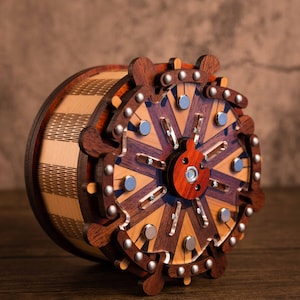 Puede incluir: Una caja de rompecabezas de madera con un dise&ntilde;o circular que presenta detalles intrincados y un mecanismo rotatorio central. La caja est&aacute; hecha de varios tonos de madera marr&oacute;n y tiene un patr&oacute;n decorativo.