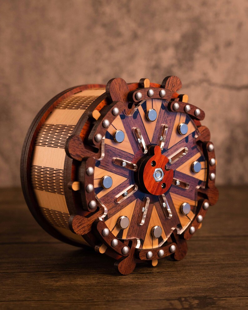 Puede incluir: Una caja de rompecabezas de madera con un dise&ntilde;o circular que presenta detalles intrincados y un mecanismo rotatorio central. La caja est&aacute; hecha de varios tonos de madera marr&oacute;n y tiene un patr&oacute;n decorativo.
