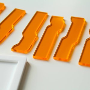 Puede incluir: Varias piezas de acrílico naranja transparente con un diseño único con muescas se disponen sobre una superficie blanca. Las piezas rectangulares presentan una hendidura curva. Un marco blanco es visible.