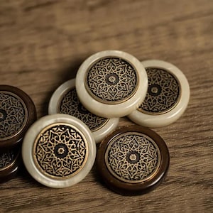 Può includere: Primo piano di sei pezzi da gioco rotondi con intricati disegni dorati e neri. I pezzi sono realizzati in materiali bianco e marrone.