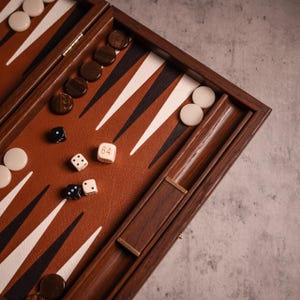 Puede incluir: Un juego de backgammon de madera con una superficie de juego de cuero marrón, con triángulos blancos y negros. El juego incluye fichas marrones y blancas y dados. Un dado muestra el número 64. El juego está abierto, listo para jugar.