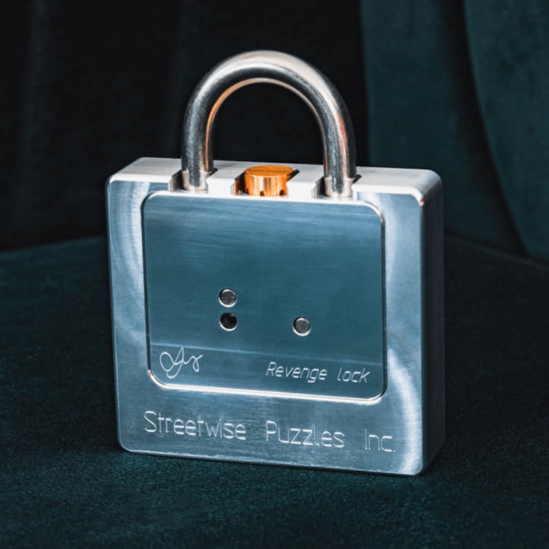 Op de afbeelding: Een zilveren metalen slot met een zilveren hangslot en een kleine oranje knop. Het slot heeft de tekst "Revenge lock" en "Streetwise Puzzles Inc." erop.