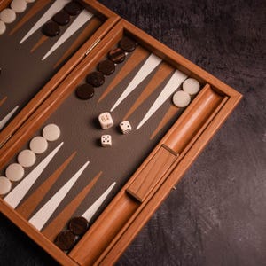 以下が含まれることがあります： 木製のバックギャモンセット。ゲーム盤は開かれており、茶色と白の三角形のパターンで、茶色と白の駒が配置されています。3つのサイコロがあり、1つは数字64を示しています。
