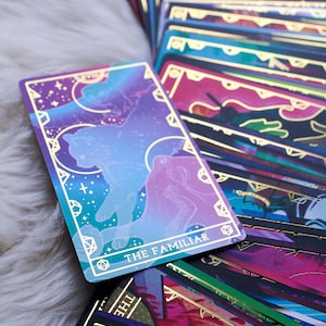 Könnte beinhalten: Ein Tarotkartenset mit einem himmlischen Thema. Die Karte "THE FAMILIAR" zeigt eine Katzensilhouette vor einem lila-blauen Nebelhintergrund. Goldakzente und geometrische Formen rahmen das Bild ein. Weitere Karten sind sichtbar.