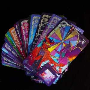 Puede incluir: Un abanico de cartas de tarot con ilustraciones vibrantes. Las cartas presentan dise&ntilde;os coloridos, incluyendo una figura de diablo con traje rojo y una rueda de la fortuna. Las cartas tienen texto japon&eacute;s y est&aacute;n sobre un fondo negro.