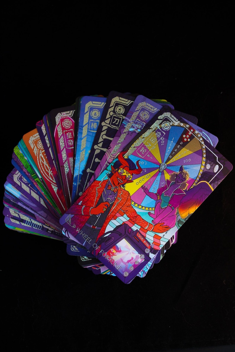 Puede incluir: Un abanico de cartas de tarot con ilustraciones vibrantes. Las cartas presentan dise&ntilde;os coloridos, incluyendo una figura de diablo con traje rojo y una rueda de la fortuna. Las cartas tienen texto japon&eacute;s y est&aacute;n sobre un fondo negro.