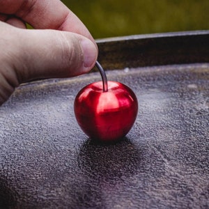 Könnte beinhalten: Eine glänzende rote Metallkirsche mit einem schwarzen Stiel. Die Kirsche liegt auf einer dunklen, strukturierten Oberfläche.