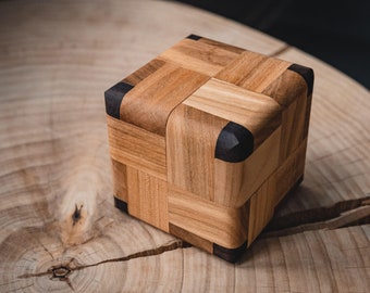May include: A wooden cube puzzle with a light brown wood finish and black accents. The cube is made of multiple pieces that fit together to form a larger cube.