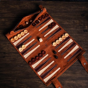 May include: A brown leather backgammon set with white and brown playing pieces. The set is rolled up and has a zipper closure. The set is on a wooden surface.