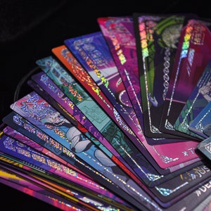 Puede incluir: Un abanico de cartas de tarot con dise&ntilde;os hologr&aacute;ficos coloridos. Las cartas presentan ilustraciones en tonos morados, azules, rosas y verdes, con bordes plateados. Las cartas est&aacute;n dispuestas sobre un fondo negro, creando una exhibici&oacute;n visualmente impactante.
