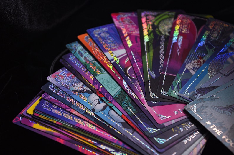 Puede incluir: Un abanico de cartas de tarot con dise&ntilde;os hologr&aacute;ficos coloridos. Las cartas presentan ilustraciones en tonos morados, azules, rosas y verdes, con bordes plateados. Las cartas est&aacute;n dispuestas sobre un fondo negro, creando una exhibici&oacute;n visualmente impactante.