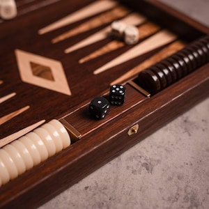 Può includere: Un gioco da tavolo backgammon in legno con pezzi avorio e marrone scuro. Due dadi neri con punti bianchi sono visibili, insieme a una pila di pezzi marrone scuro. Il tabellone ha un intarsio a forma di diamante.