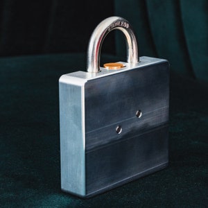 Op de afbeelding: Een zilveren metalen slot met een roestvrijstalen beugel. Het slot heeft een vierkant lichaam en een klein, rond sleutelgat. Het slot is gesloten en de beugel is vastgemaakt.