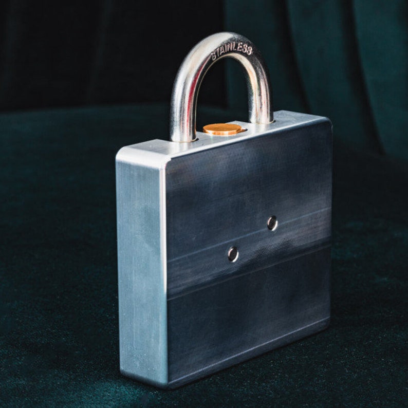 Op de afbeelding: Een zilveren metalen slot met een roestvrijstalen beugel. Het slot heeft een vierkant lichaam en een klein, rond sleutelgat. Het slot is gesloten en de beugel is vastgemaakt.