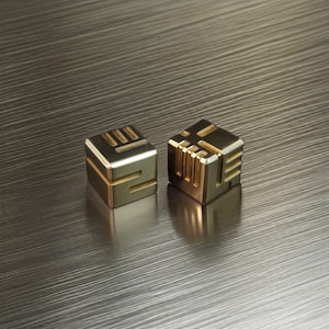 May include: Two small, gold-coloured metal cubes with geometric designs. Each cube has a different pattern of lines and shapes etched into its surfaces. The cubes are displayed on a textured, metallic surface, creating a modern and minimalist aesthetic.