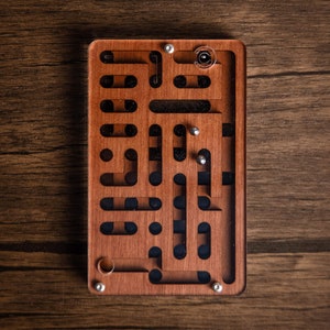 Puede incluir: Un juego de laberinto rectangular de madera con una tapa de acrílico transparente. El laberinto presenta una serie de caminos interconectados y pequeñas bolas de metal. La madera tiene un tono marrón rojizo cálido, sobre un fondo de vetas oscuras.