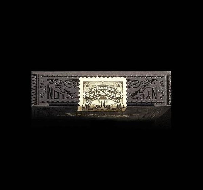 Op de afbeelding: Een zwarte speelkaartenbox met gouden accenten. De doos heeft de woorden "Not" en "NYC" aan de voorkant en een gouden stempel met de woorden "Stranger Strange" en "14" erop.