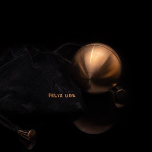 May include: A gold-colored metal sphere with a smooth, shiny surface. The sphere is resting on a black velvet pouch with the text "FELIX URE" embroidered on it.
