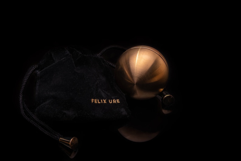 May include: A gold-colored metal sphere with a smooth, shiny surface. The sphere is resting on a black velvet pouch with the text "FELIX URE" embroidered on it.