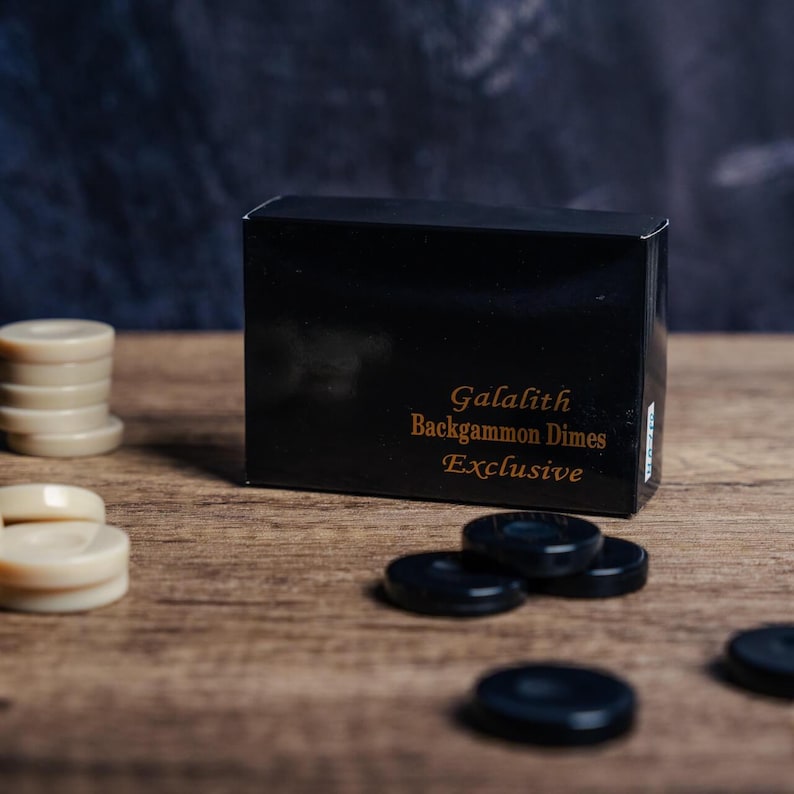 Puede incluir: Una caja negra con el texto "Galalith Backgammon Dimes Exclusive" en ella. La caja est&aacute; sobre una superficie de madera con piezas de juego negras y blancas esparcidas a su alrededor.