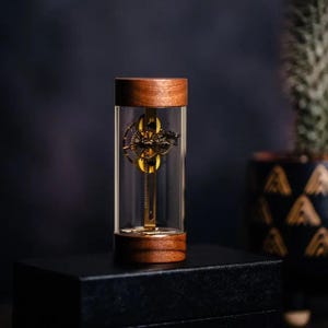 May include: A small, intricate clockwork mechanism encased in a clear glass cylinder with a walnut wood base. The clockwork is made of brass and has a gold finish.