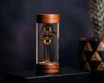 May include: A small, intricate clockwork mechanism encased in a clear glass cylinder with a walnut wood base. The clockwork is made of brass and has a gold finish.