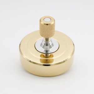 Può includere: Una trottola in metallo color oro con una base circolare liscia e uno stelo verticale centrale. Lo stelo presenta una superficie di presa zigrinata e testurizzata e un piccolo inserto circolare piatto color argento sulla parte superiore.