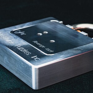 Op de afbeelding: Een zilveren metalen slot met de tekst "Revenge Lock" en "Puzzle Wise Inc." gegraveerd aan de zijkant. Het slot heeft drie kleine gaten aan de bovenkant.