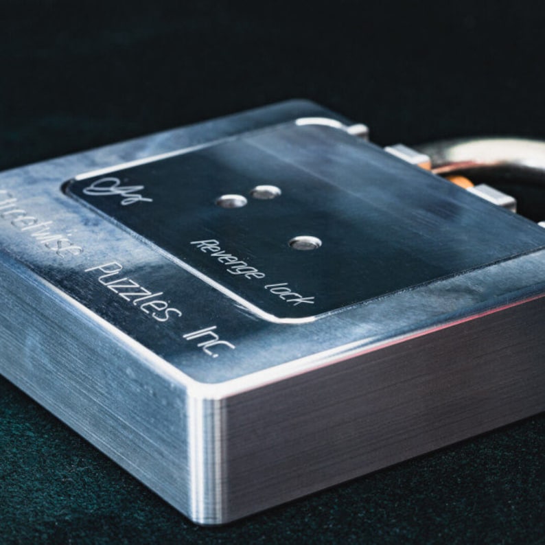 Op de afbeelding: Een zilveren metalen slot met de tekst "Revenge Lock" en "Puzzle Wise Inc." gegraveerd aan de zijkant. Het slot heeft drie kleine gaten aan de bovenkant.