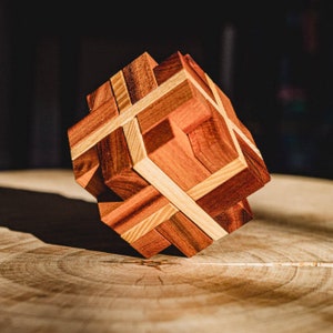 May include: A wooden puzzle cube with interlocking pieces in shades of brown and tan. The cube is constructed from various wood grains, creating a geometric design. The puzzle sits on a wooden surface, with natural light casting shadows.