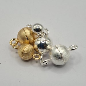 Könnte beinhalten: Ein Satz magnetischer Verschlüsse für die Schmuckherstellung. Die Verschlüsse sind aus Silber- und Goldmetall gefertigt und haben eine gebürstete Oberfläche. Die Verschlüsse sind rund und haben einen Durchmesser von etwa 1,5 cm.