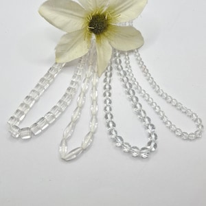 Op de afbeelding: Vier strengen van heldere glazen kralen, twee met vierkante kralen, een met ovale kralen en een met ronde kralen.