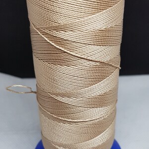 Pu&ograve; includere: Un'bobina di filo beige avvolta su una bobina di plastica blu. Il filo &egrave; un materiale intrecciato, testurizzato e lucido.
