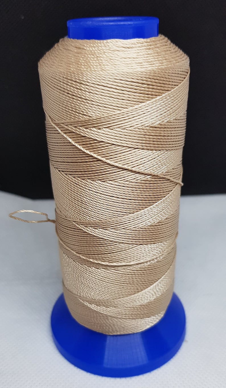 Pu&ograve; includere: Un'bobina di filo beige avvolta su una bobina di plastica blu. Il filo &egrave; un materiale intrecciato, testurizzato e lucido.