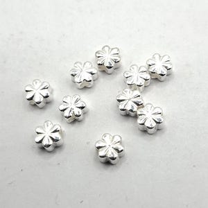Könnte beinhalten: Eine Ansammlung kleiner, silberfarbener, blütenförmiger Perlen. Jede Perle hat ein detailliertes Blütenblattdesign und eine leicht abgerundete Form. Die Perlen sind auf einer weißen Oberfläche verstreut, was eine zarte und elegante Ästhetik erzeugt.