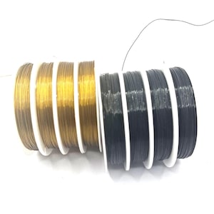 Könnte beinhalten: Spulen mit Basteldraht in Gold, Dunkelgrau und Schwarz. Der Draht ist auf weißen Kunststoffspulen aufgewickelt. Der goldene Draht hat einen metallischen Glanz, während die dunkelgrauen und schwarzen Drähte matt sind. Das Bild ist vor weißem Hintergrund.