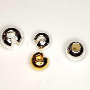 Könnte beinhalten: Eine Sammlung von vier Schmuckzubehörteilen, bestehend aus drei silberfarbenen Quetschperlenkappen mit C-förmiger Öffnung und einer goldfarbenen Quetschperlenkappe, sowie einer glatten, kugelförmigen silberfarbenen Perle mit einem Mittelloch.