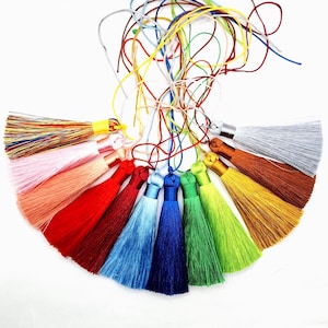 Könnte beinhalten: Eine Sammlung bunter Quasten in verschiedenen Rot-, Rosa-, Blau-, Grün-, Gelb- und Brauntönen. Jede Quaste hat eine passende Kordel zum Aufhängen. Die Quasten sind in einem Halbkreis auf weißem Hintergrund angeordnet.