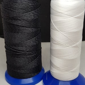 Pu&ograve; includere: Due bobine di filo, una nera e una bianca, sono avvolte su bobine di plastica blu. Le bobine sono su una superficie bianca.
