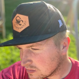 May include: A black and brown trucker hat with a scorpion logo and the text "Scorpio Knives". The hat has a mesh back and a white B logo on the side.