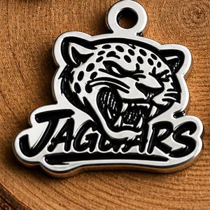 May include: A silver-colored pendant featuring a detailed jaguar head with the word "JAGUARS" in bold, black lettering. The jaguar has a fierce expression, with visible teeth and spotted markings. The pendant has a loop at the top for attachment.