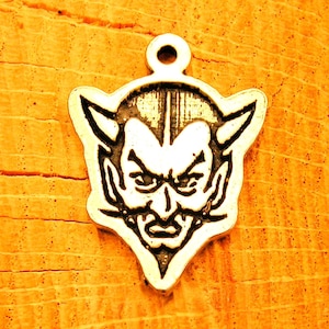 May include: A silver charm in the shape of a devil's head with horns, a pointed nose, and a frowning expression.